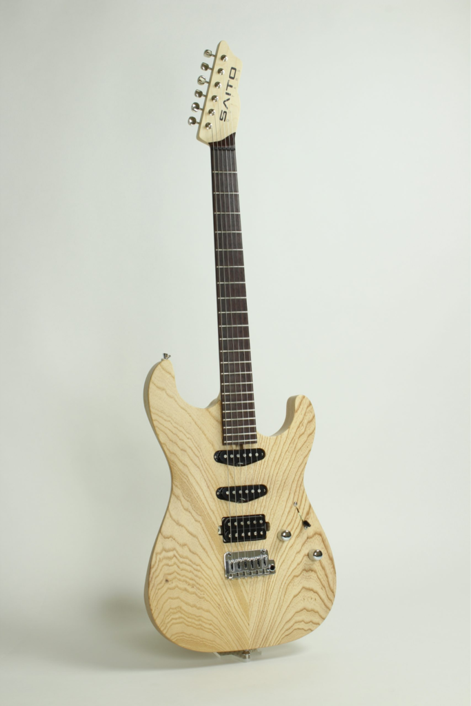 Saito Guitars electric guitar with natural ash body, visible wood grain, HSS pickup configuration, fixed bridge, and minimalist control layout on a light background.